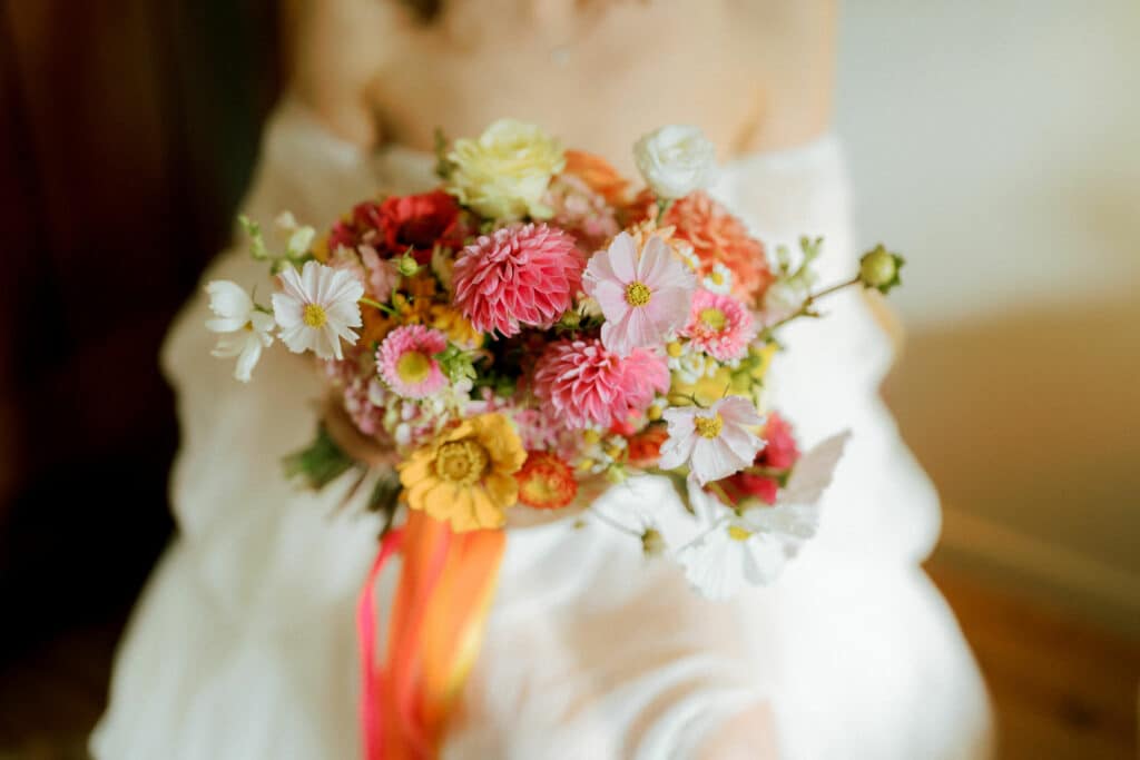 Bride holding her bridal bouquet made of pale pink, hot pink, orange, coral, yellow, salmon and white flowers. The bouquet contains cosmos, dahlias, strawflowers, zinnias, lisianthus roses and hydrangea.