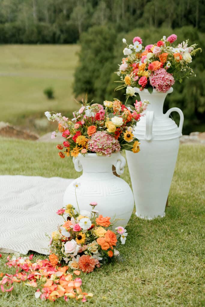 2 white urns and floor arrangement of bright flowers for wedding ceremony at the side of the ceremony space outdoors