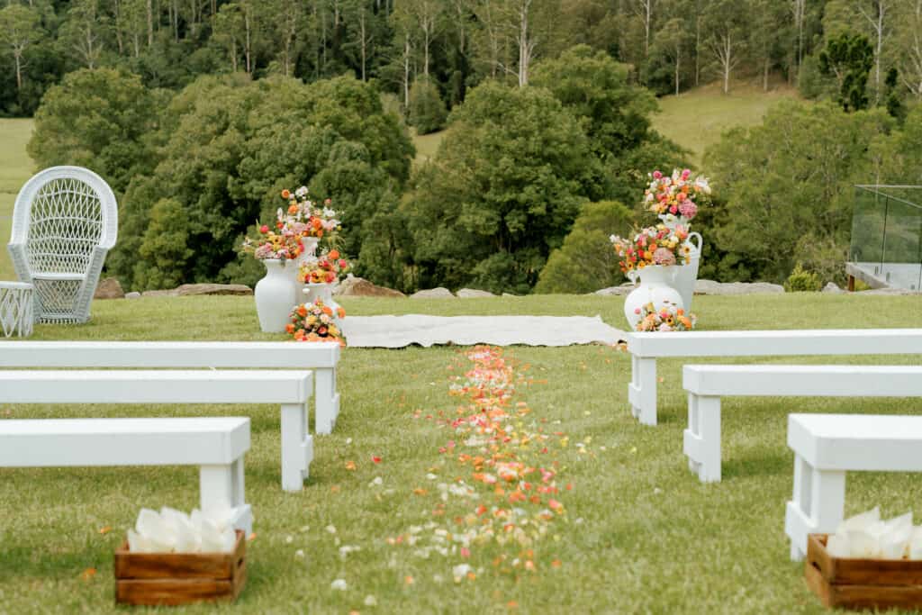 Wedding venue set up with white benchs on either side of an aisle with a rug and white urns filled with flowers in front of the benches. Flower petals are spread on the grass down the aisle