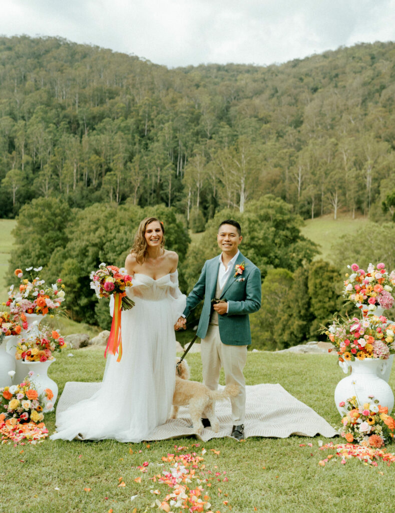 Bride and Groom standing on a rug with their dog at an outdoor wedding with trees and a valley behind them. Groom is wearing a green jacket and vest with flowers on his lapel. Bride is wearing a long wedding gown holding a bridal bouquet with bright floating ribbon in orange, pink and gold. There are white urns of summery flowers in colours of orange, pink, yellow and white on both sides of the rug and flower petals on the grass in front of the smiling couple