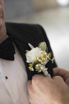 A Groom's Boutonneire with White rose and daisies being pinned to a black suit