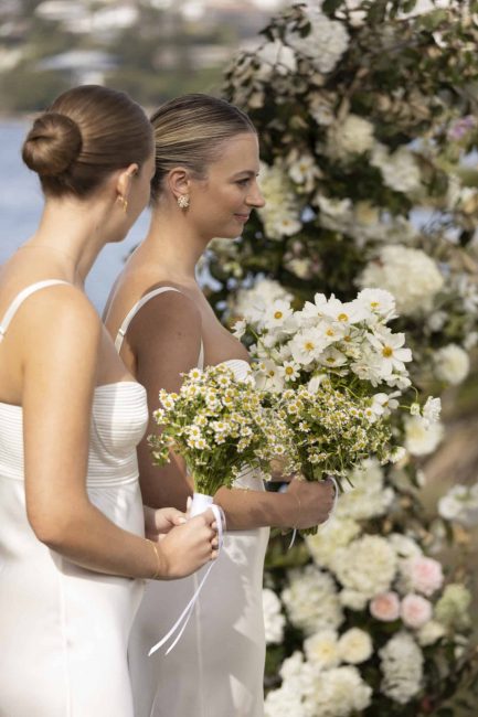 Two bridesmaids during the wedding ceremony wearing white dresses holding white daisy bouquets with asymmetrical flower towers in the background.